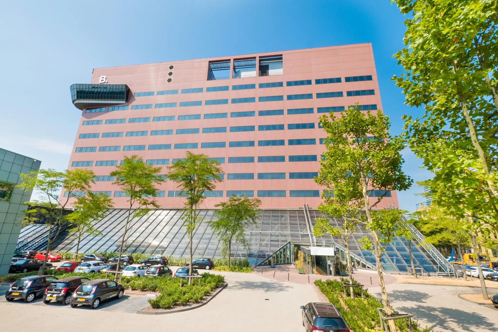 Modern kantoorgebouw aan de Johan Huizingalaan in Amsterdam met parkeerplaatsen en glazen entree, gezien op een zonnige dag.